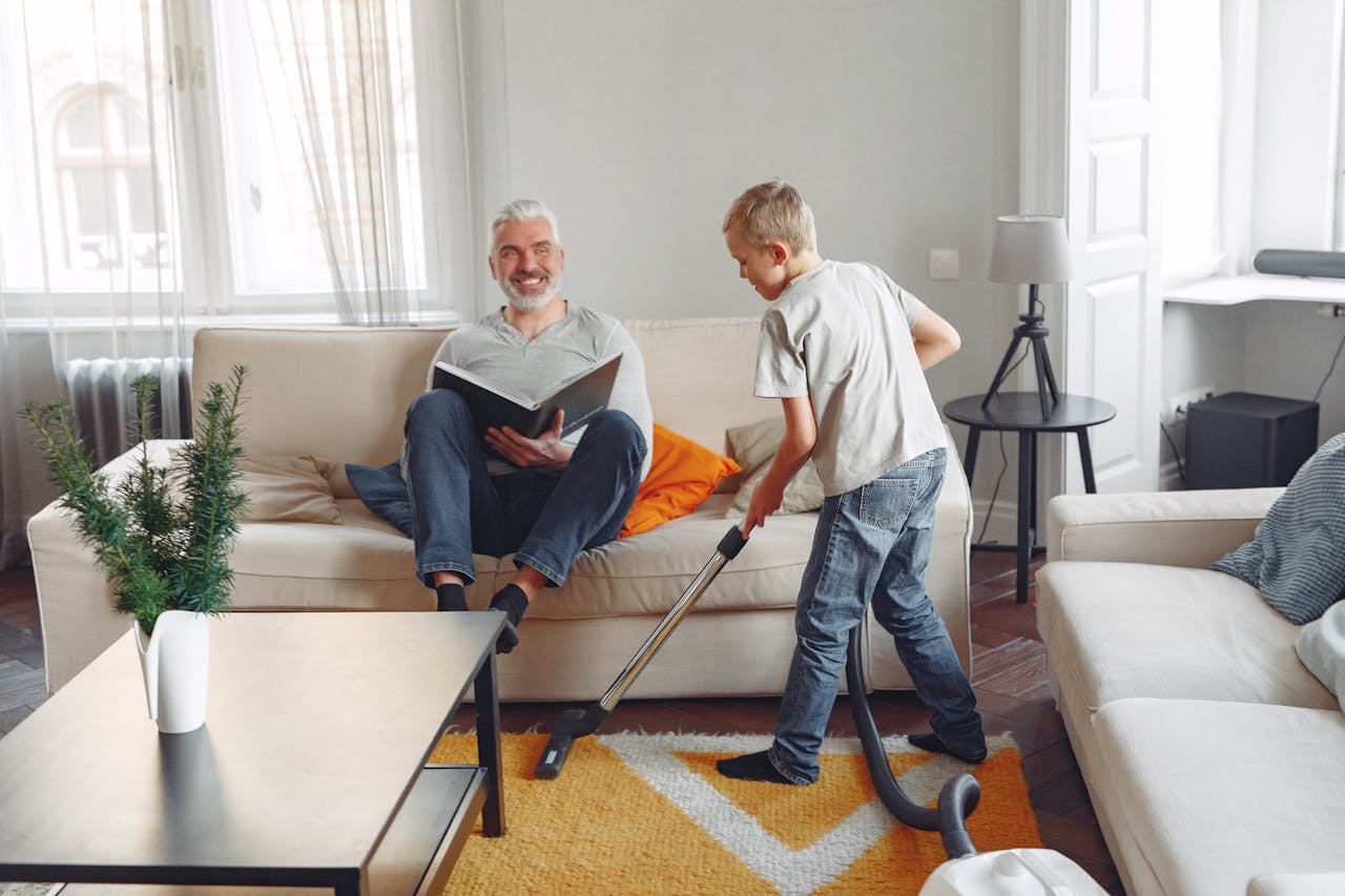 services-02 A father relaxing while his son vacuums in a cozy living room setting, showcasing family bonding.